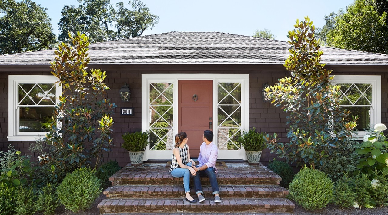 A millennial couple sits on the front steps of their home and turns to face the door.